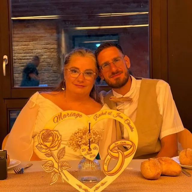 Rachel et Jean-Charles le jour de leur mariage, devant un cœur en bois gravé à leurs prénoms
